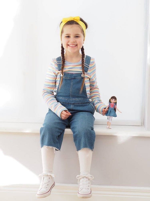 Girl holding Lottie Doll with Dark brown hair. in white leggings and jean skirt and red/white stripe shirt